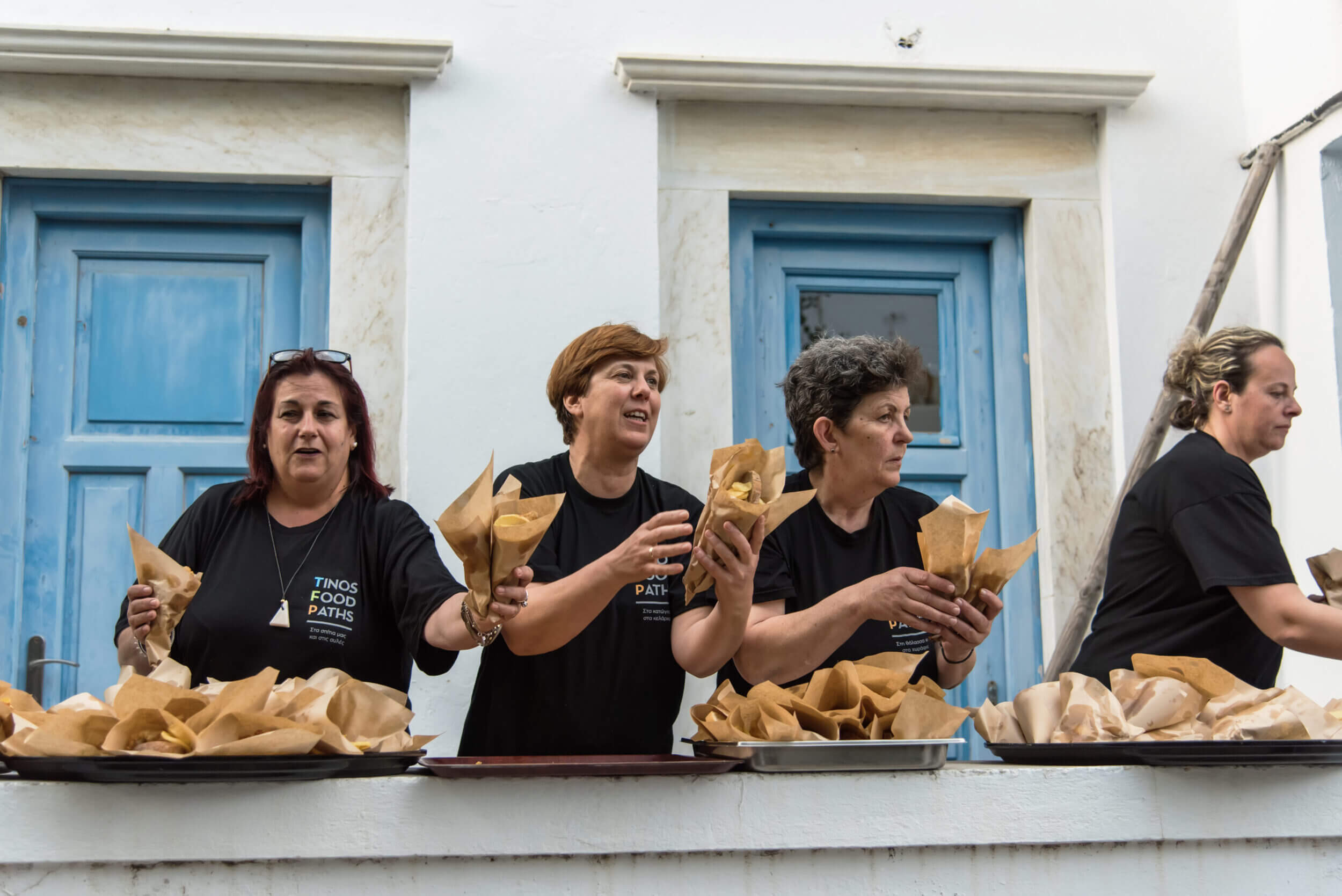 Γυναίκες με μπλουζάκια Tinos Food Paths μοιράζουν φαγητό σε χάρτινα χωνάκια